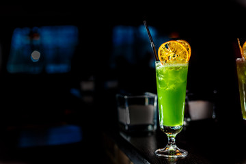 A green cocktail in a tall glass stands on the counter of a dark bar.