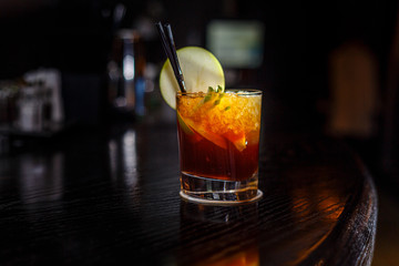 A cocktail with whiskey or cognac or Cola in a highball glass is on the counter of a dark bar.