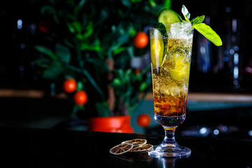 A green cocktail or lemonade with cucumber in a tall glass stands on the counter of a dark bar.