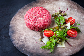 Gourmet tartar raw from beef fillet with lettuce, tomatoes and parmesan with capers as closeup on a rustic modern design plate with copy space left