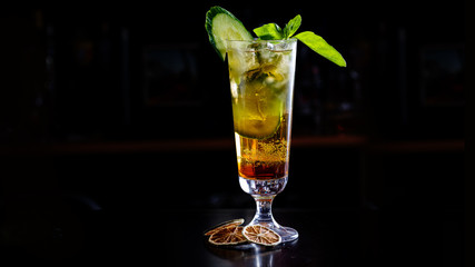 A green cocktail or lemonade with cucumber in a tall glass stands on the counter of a dark bar.