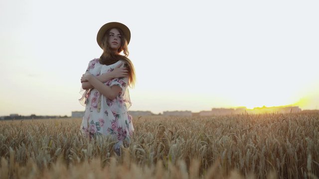 Portrait of young faraway lady in a hat, stands in poses among field on sunset