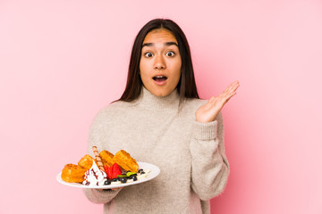 Young asian woman holding a waffle isolated surprised and shocked.