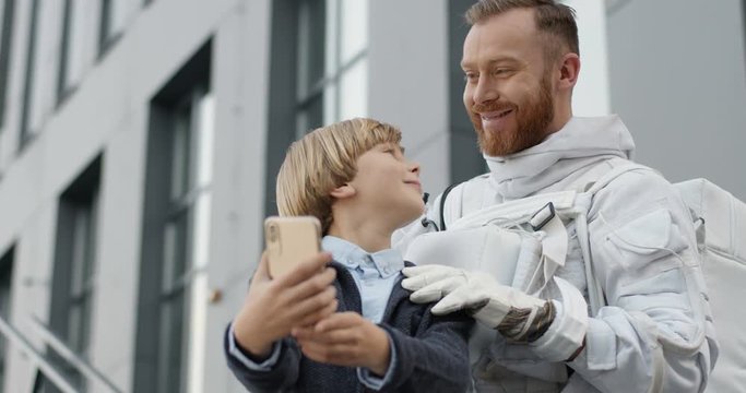 Cute Caucasian small boy taking selfie photo with his handsome father in space suit on smartphone camera. Spaceman coming back home to little son and posing to phone photos.