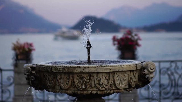 Slow motion. Glide shot. Fountain on promenade in Menaggio Italy. Lake Como. Alp mountains. Summer luxury tourism landmark & romantic honeymoon travel destination.