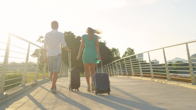 SLOW MOTION, LENS FLARE, COPY SPACE: Young newlyweds carry their suitcases across an overpass and to the airport at sunset. Golden evening sun rays shine on tourist couple on their way to the airport.