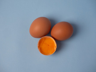 Two whole raw brown chicken eggs and a broken half egg with yellow yolk closeup on a blue background top view.