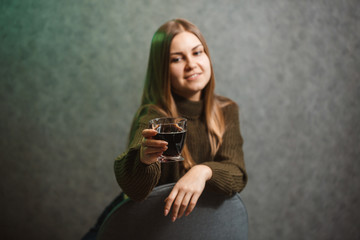 A girl with a glass with black liquid sits on a chair on a gray background