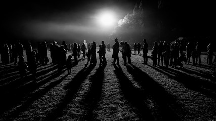 Black and white picture of a people standing one after another in front of bright light and casting long shadows on a grass on a local festival smoothed by a motion.