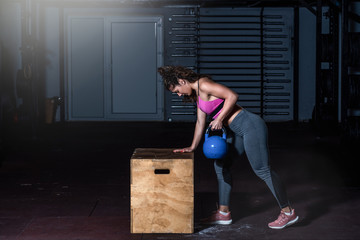 Young sweaty fit muscular strong girl doing hardcore cross workout training for back muscles with heavy kettlebell on the wooden jump box in the gym dark image real people exercising