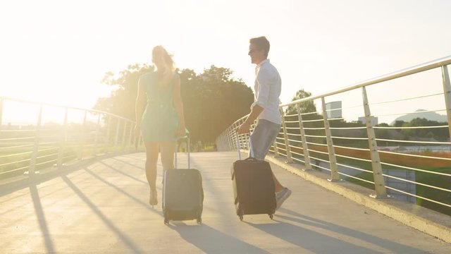 SLOW MOTION, LENS FLARE: Man excited to go on trip jumps in the air and pumps his fist while walking to the airport with girlfriend at sunset. Young couple is excited to go on their summer vacation.