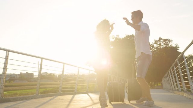 SLOW MOTION, LENS FLARE, LOW ANGLE: Happy newlywed couple celebrate going on honeymoon while walking to airport with their baggage. Man high fives with girlfriend while walking to airport at sunset.