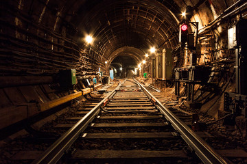 Brightly lit track development. Railroad tracks pass straight and turn left. Red traffic lights light up. Vertical iron boxes are visible. Electric wires run along the walls. 