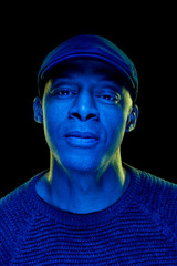 Black man with blue and yellow light, wearing a flat cap, isolated on black background, looking at the camera. Confidence concept. Vertical