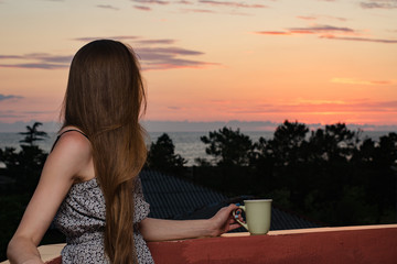 Fototapeta premium Young woman with a cup in her hand looking at sea and sunset. Side view