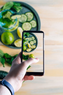 ands picking up a smartphone taking a picture of a refreshing water with cucumber, lime and good herb. Healthy drink and detoxification