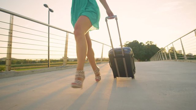 SLOW MOTION, LENS FLARE, CLOSE UP, LOW ANGLE: Cinematic shot of woman rushing to the airport on a sunny evening. Girl in heels sprints with her luggage to catch her flight to her vacation destination
