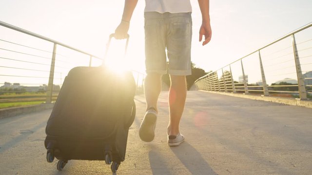 SLOW MOTION, LENS FLARE, CLOSE UP, LOW ANGLE: Unrecognizable man in shorts walks across an overpass with his luggage. Young male digital nomad drags his suitcase to the airport to move someplace else.
