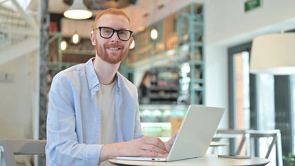 Redhead Man with Laptop Smiling at Camera in Cafe