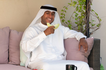 Happy, handsome Emirati man wearing traditional kandura or thobe  & ghutra sitting outdoors  eating...
