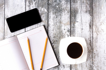Smartphone with black screen on a rough wooden background with cup of black coffee. An opened notebook with two pencils on it lie next to them.