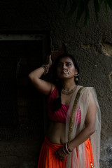 Fototapeta premium Young and attractive Indian Bengali brunette woman standing in front of a vintage house window wearing Indian traditional ethnic cloths pink and orange skirt blouse. Indian lifestyle and fashion