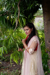 Young and beautiful Indian Bengali brunette woman standing thoughtfully in green nature below a tree wearing Indian traditional ethnic white and red gown. Indian lifestyle and fashion