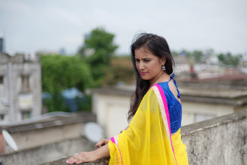 Young and beautiful Indian Bengali brunette woman in Indian traditional dress yellow sari and blue blouse is standing thoughtfully leaning on rooftop wall under blue sky with clouds. Indian lifestyle