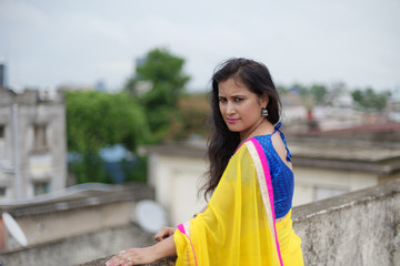 Young and beautiful Indian Bengali brunette woman in Indian traditional dress yellow sari and blue blouse is standing thoughtfully leaning on rooftop wall under blue sky with clouds. Indian lifestyle