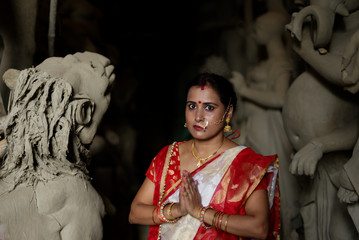 Portrait of an young and beautiful Indian Bengali brunette woman in red and white traditional ethnic sari in front of the clay idol of Hindu Bengali goddess Durga. Indian culture, religion and fashion