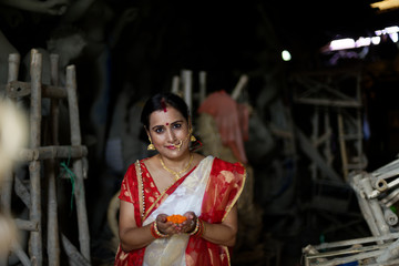 Fototapeta na wymiar An young and beautiful Indian Bengali brunette woman in red white traditional ethnic sari holding a puja thali for worshiping goddess Durga clay idol . Indian culture, religion and fashion