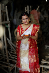 Obraz premium An young and beautiful Indian Bengali brunette woman in red white traditional ethnic sari holding a puja thali for worshiping goddess Durga clay idol . Indian culture, religion and fashion