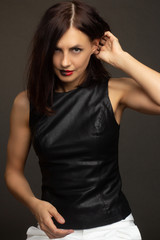 a brunette in a black leather sleeveless blouse and white trousers poses on a gray background. warm toning. lips painted black and red