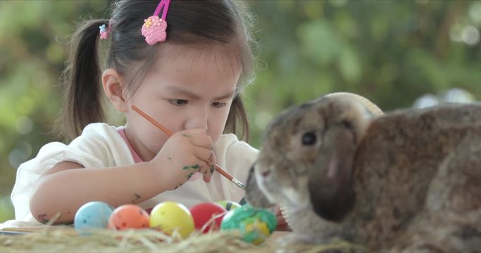 Close up of asian little girl coloring easter eggs with colors and brush near a brown rabbit on easter festive desk. Preparation of the Easter. Slow motion