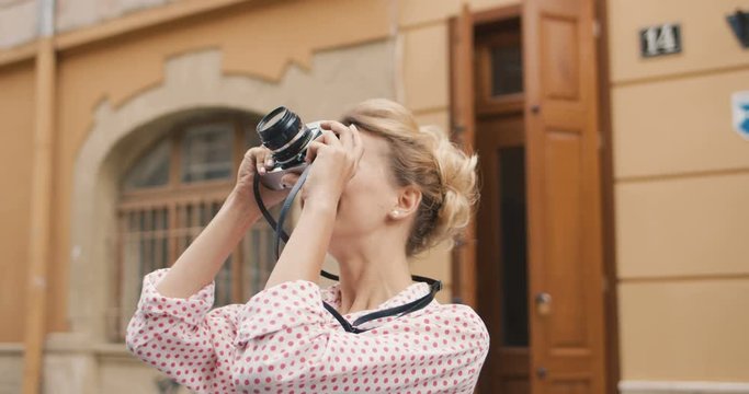 Portrait of vintage good looking Caucasian blonde woman taking photos on retro camera at daytime while walking down the street. Female beautiful trendy tourist making photos outside. Glamour concept