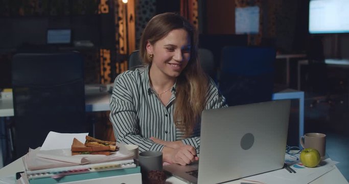 Young woman having video call with boss on her laptop in night office. Angry female employee showing fuck you after video conversation while sitting at her desk in workplace.