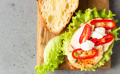 Fresh croissant sandwich with grilled chicken or turkey, bell pepper, sauce and salad leaves on wooden cutting board. Breakfast.