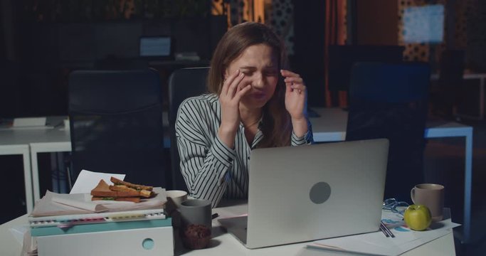 Young stressed woman looking tired and burnout while working on laptop at night office. Exhausted girl having panic because of difficulties and pressure while sitting at desk.