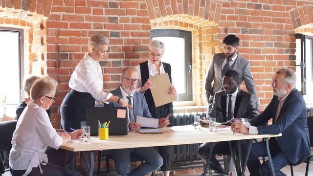 Group of businessmen create new product in market. In centre at table caucasian old male lider wear compares plans. Colleagues near give ideas