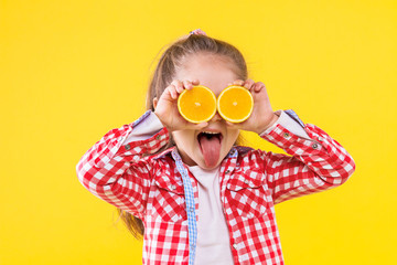 Pretty beautiful fair haired girl in checkered pink shirt is smiling on bright yellow background. Fashionable cute child is holding half of oranges like glasses. Emotional portrait concept.
