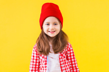 Cheerful stylish pretty little school girl dressed in pink checkered shirt, red cap, hat on yellow orange background. Cute child pupil is smiling and having fun. Emotional portrait concept.