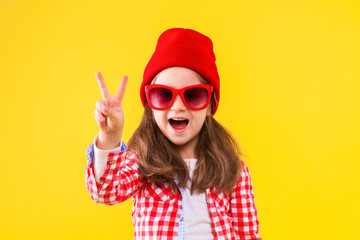 Cheerful stylish little girl dressed in pink checkered shirt, red cap, sunglasses, jeans on yellow background. Smiling cute child is showing with fingers peace sign, number two. Emotional portrait.