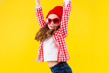 Cheerful stylish little girl dressed in pink checkered shirt, red cap, sunglasses, jeans is jumping on yellow background. Smiling cute child is dancing, having fun. Emotional portrait concept.