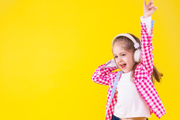Pretty cheerful fair haired girl in pink checkered shirt is listening music and dancing. Cute child in white headphones is smiling, having fun on orange yellow background. Modern children concept.