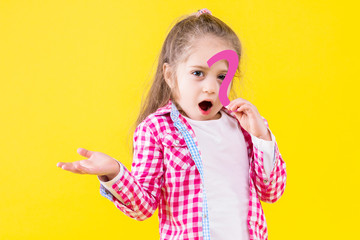 Pretty beautiful perplexed fair haired girl dressed in pink checkered shirt is holding in hand paper question sign. Cute child is smiling on yellow orange background. Emotional portrait concept.