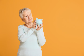 Happy senior woman with piggy bank on color background
