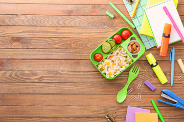 School lunch box with tasty food and stationery on wooden background