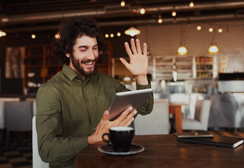 Casual bearded man waving his hand while talking through video-chat in smartphone while relaxing in cafe