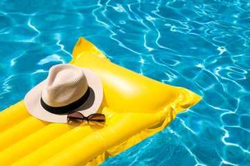 Sun hat and sunglasses resting on bright yellow inflatable raft floating in blue swimming pool - obrazy, fototapety, plakaty