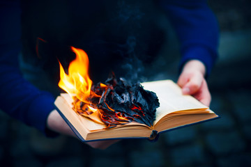 A man holds an open Bible in his hands, the pages of which burn with a bright flame. Hatred of religion. Antichrist. Hooliganism. - obrazy, fototapety, plakaty